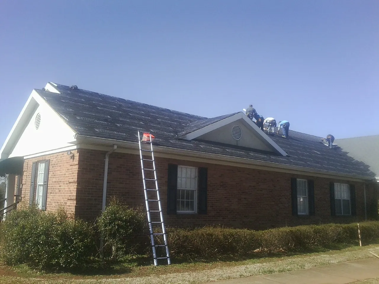 Crew tearing shingles off a brick ranch roof with a ladder set — Emergency Roof Repair work by our crew in Sound Beach