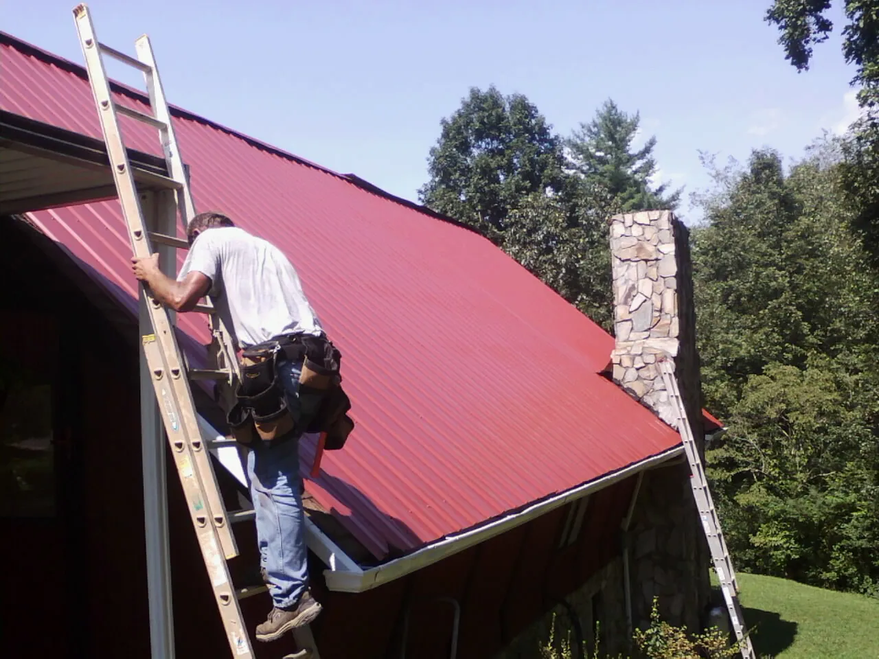 Roofer on a ladder beside a red metal roof and stone chimney — Soffit & Fascia Repair by Roofing Brothers in Sound Beach